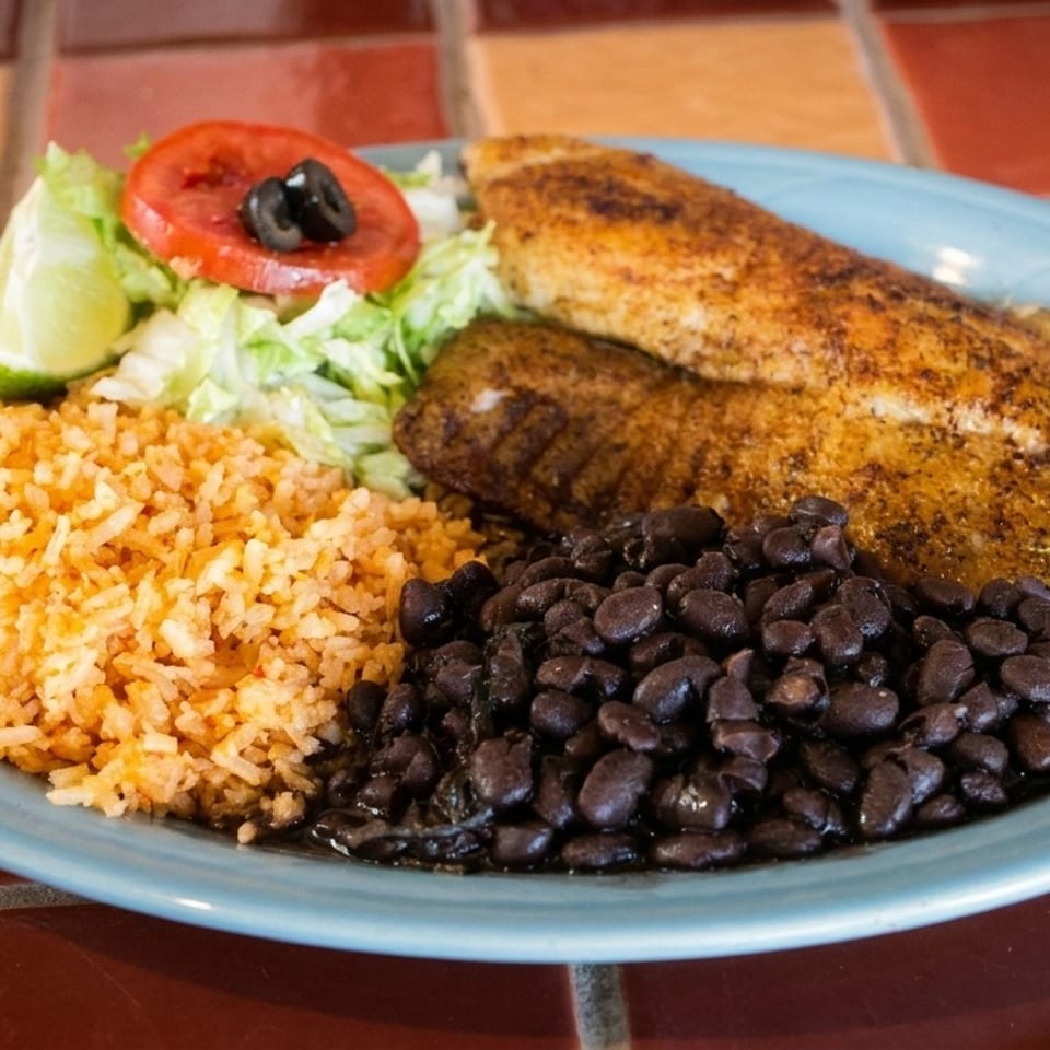 Two seasoned fish fillets, rice, black beans, and a small salad with lime on a blue plate.