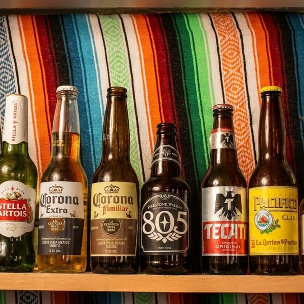 A colorful serape backdrop behind a shelf displaying various beer bottles, including Mexican brands.